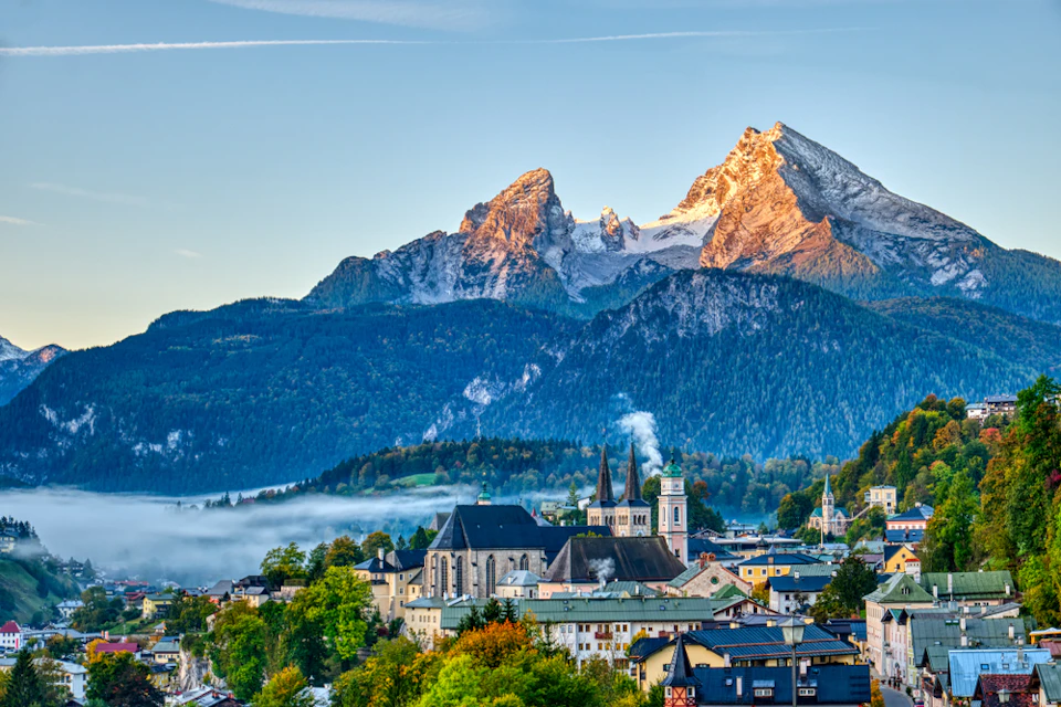 A picturesque village with a prominent church set against a backdrop of sunlit, snow-capped mountains and lush green hills.