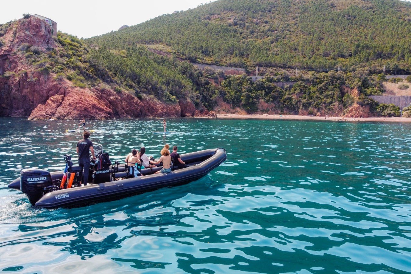 Skupina lidí na motorovém člunu v průzračné modré vodě poblíž zalesněného kopce a skalnatého pobřeží.