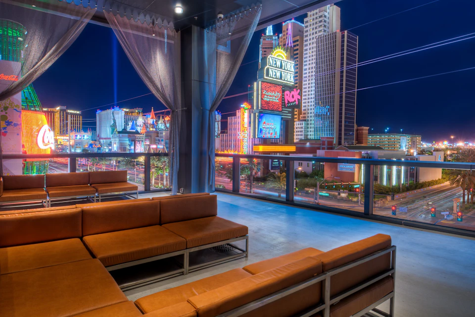 Rooftop lounge with brown couches, overlooking illuminated New York-New York hotel and casino, and other buildings at night.