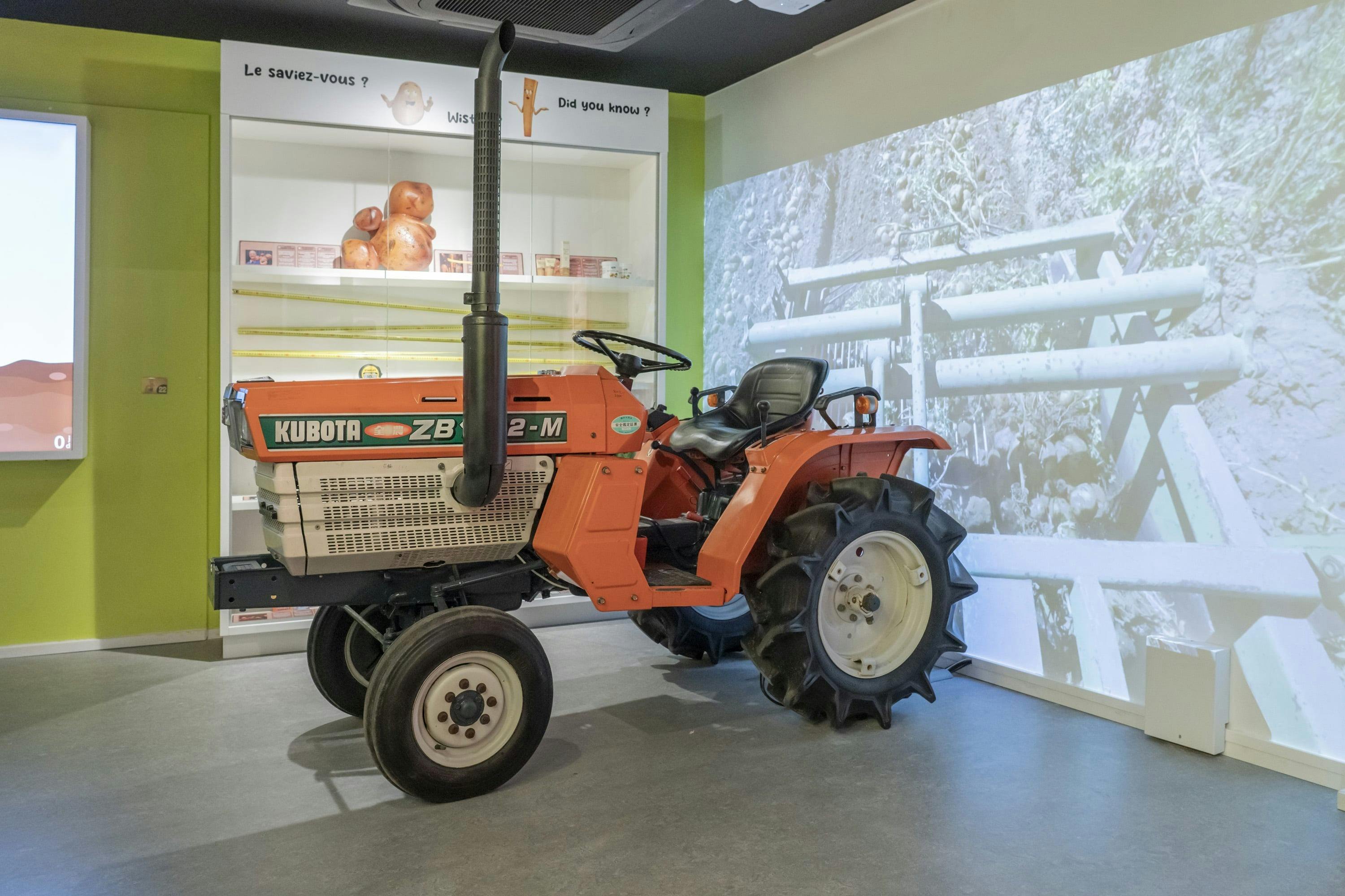 Un trattore Kubota arancione esposto all'interno, con un'immagine proiettata di una scena agricola sulla parete dietro di esso.