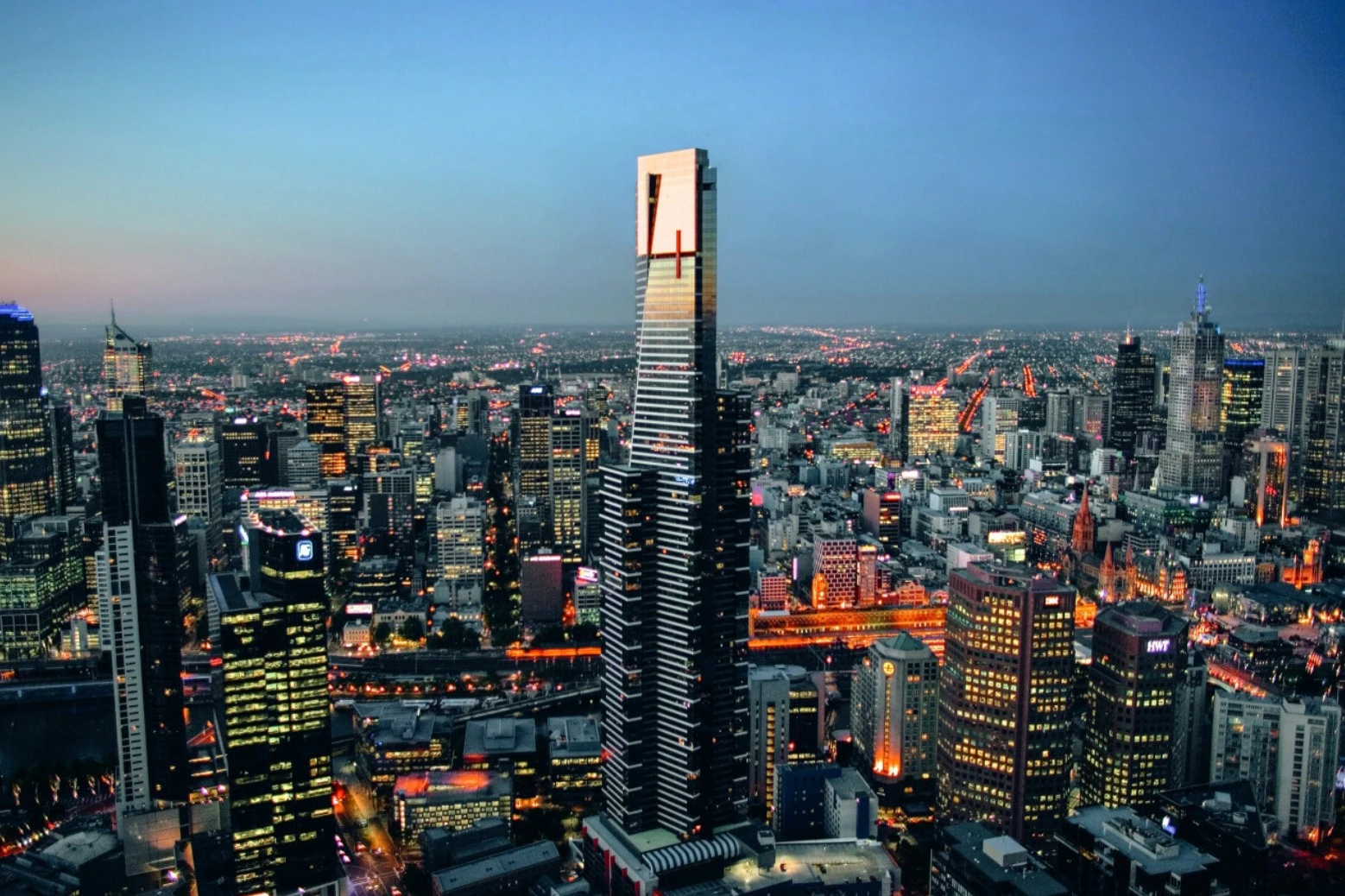 A tall skyscraper with illuminated windows in an urban cityscape during twilight, surrounded by other buildings.