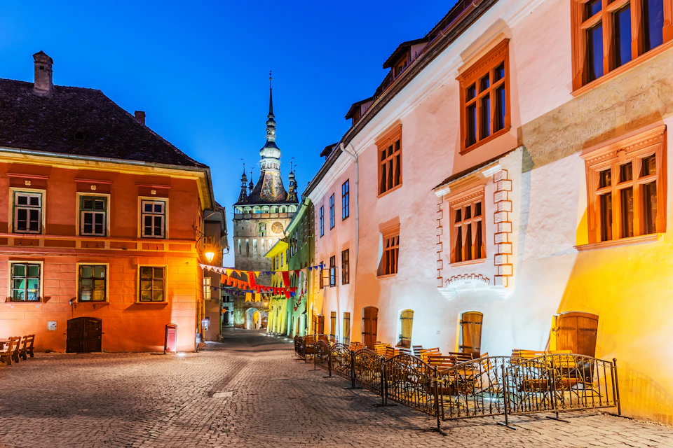 A cobblestone street lined with colorful buildings, outdoor seating, and a distant clock tower, illuminated at twilight.