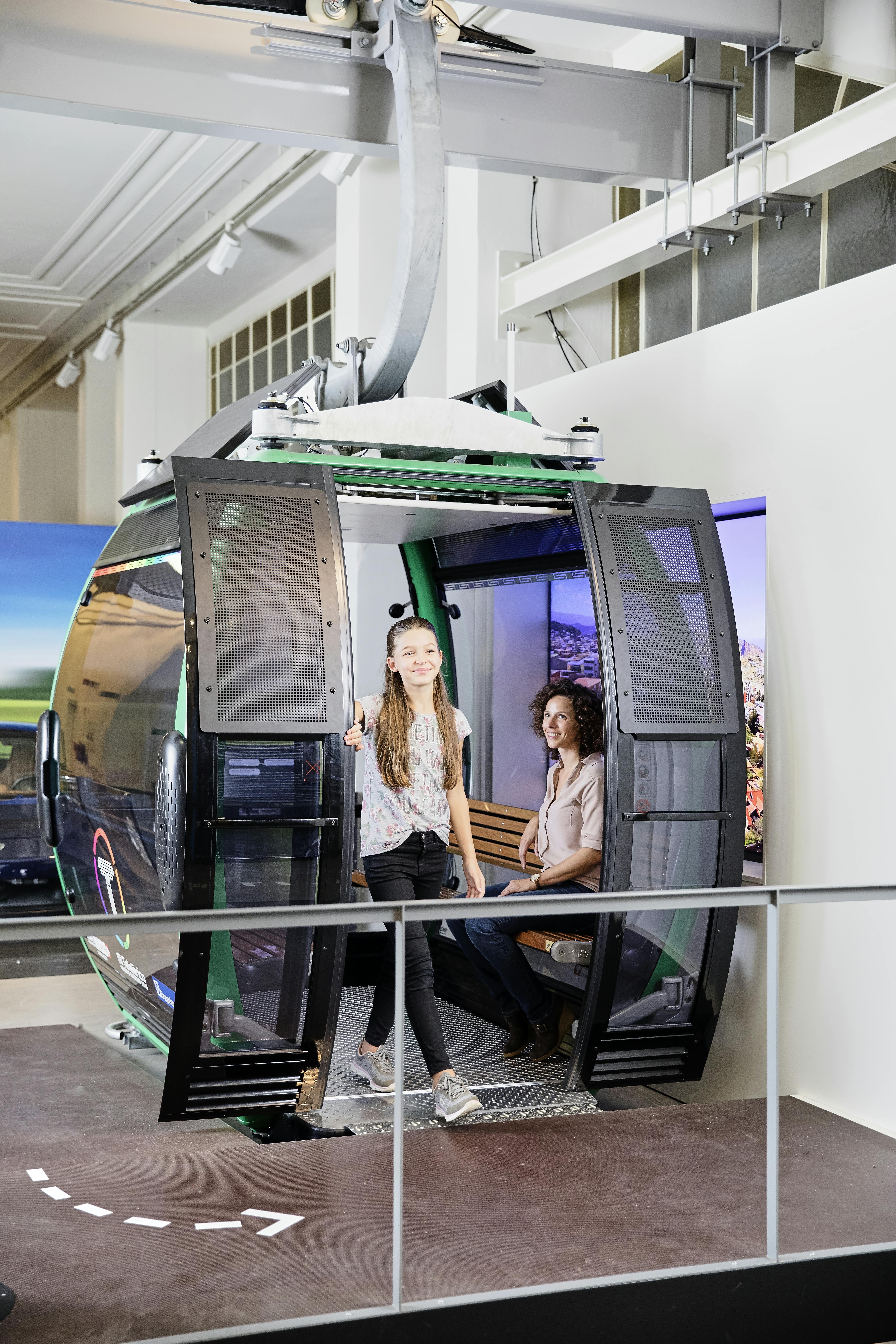 Dívka se usmívá, když vystupuje ze stacionární kabiny kabinkové lanovky v moderním výstavním prostoru se sedící ženou uvnitř.