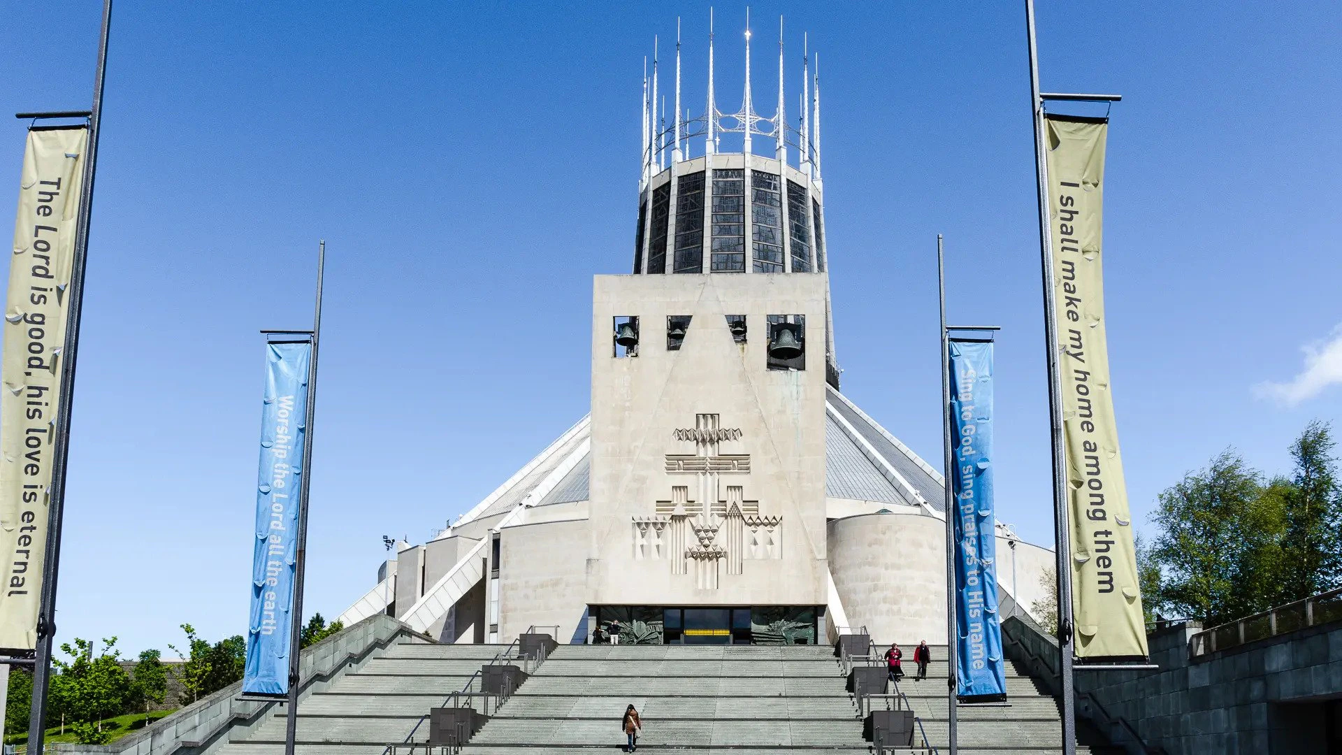 Modern cathedral with a distinctive central tower, large staircase in front, and blue banners on either side. Clear blue sky.
