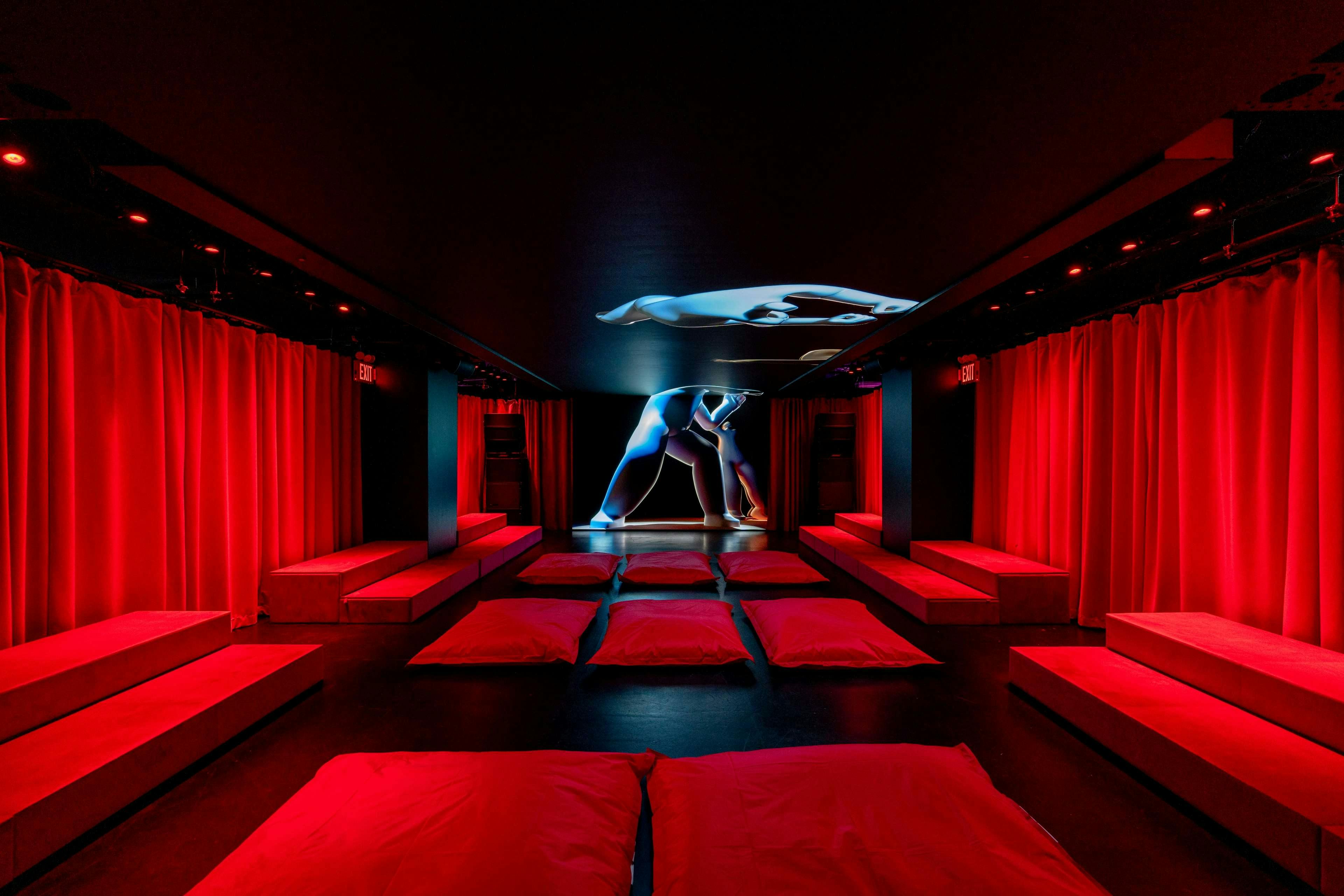 A dimly lit room with red curtains and floor cushions, a sculpture of two figures on the ceiling, and a stage at the back.