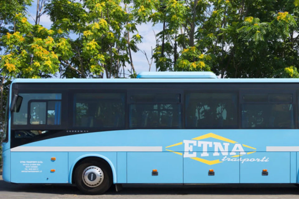 A blue bus with the words "ETNA" displayed on its side, parked in front of a row of green trees.
