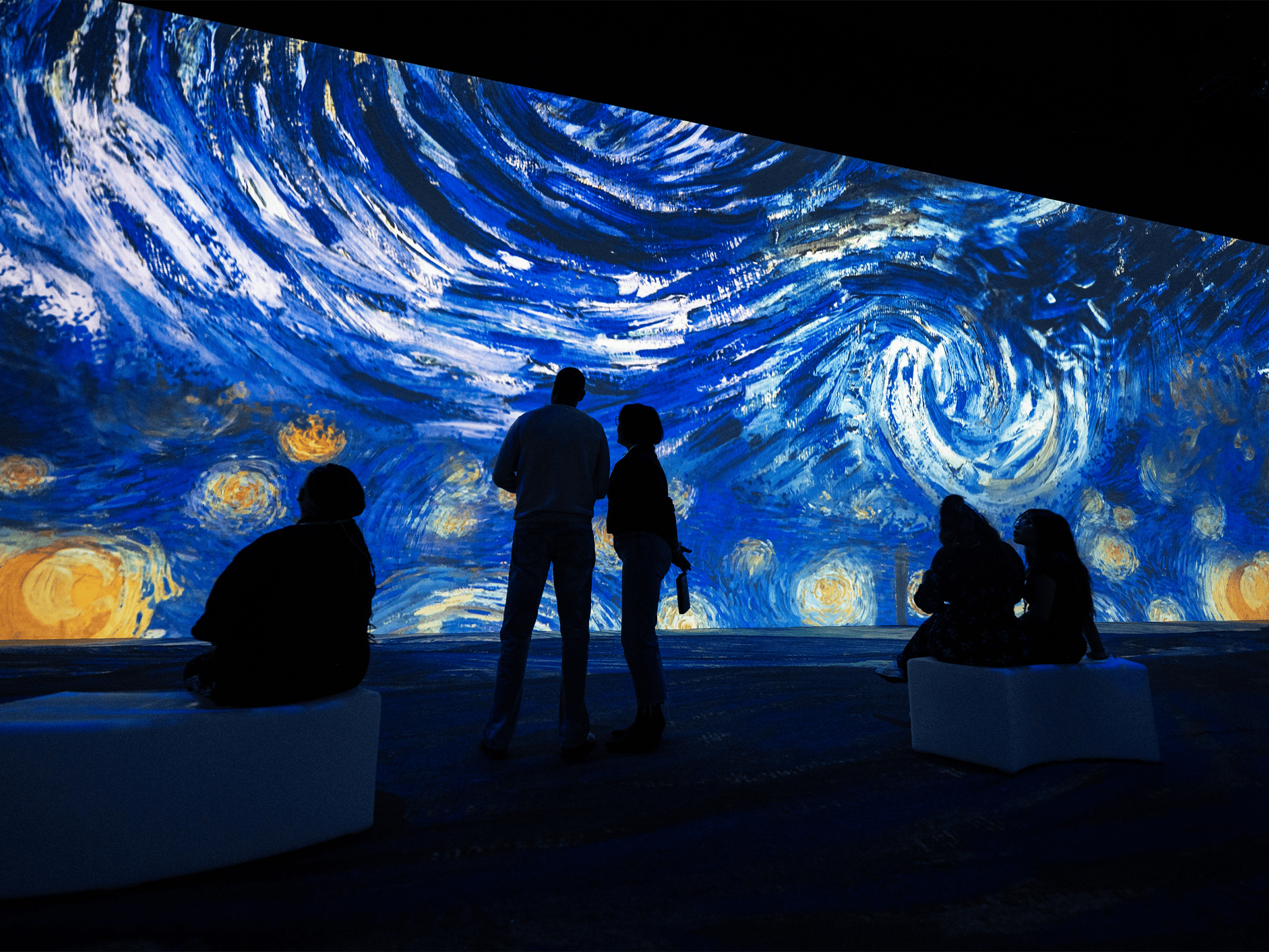 人们坐在白色长凳上，站在黑暗的房间里观看梵高《星夜》的大型灯光展览。