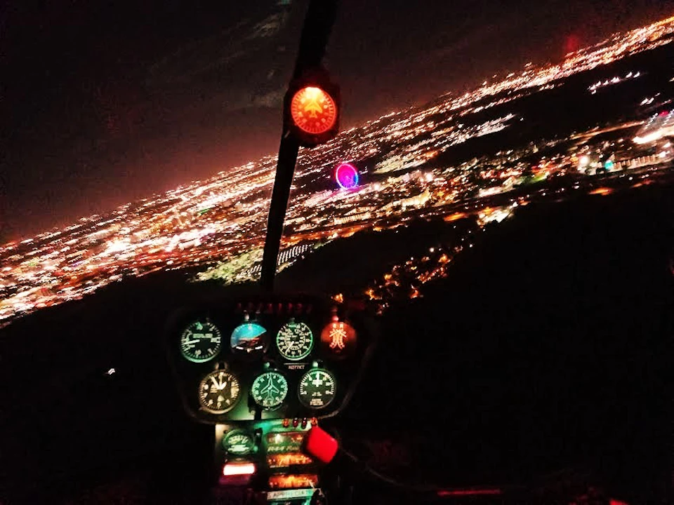 Nighttime helicopter cockpit view with illuminated instrument panel, showing a sprawling cityscape below with a prominent Ferris wheel.