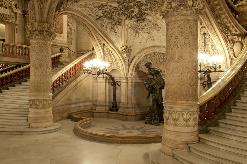 Ornate indoor staircase with detailed carvings, a large statue, and decorative lighting in a grand architectural setting.