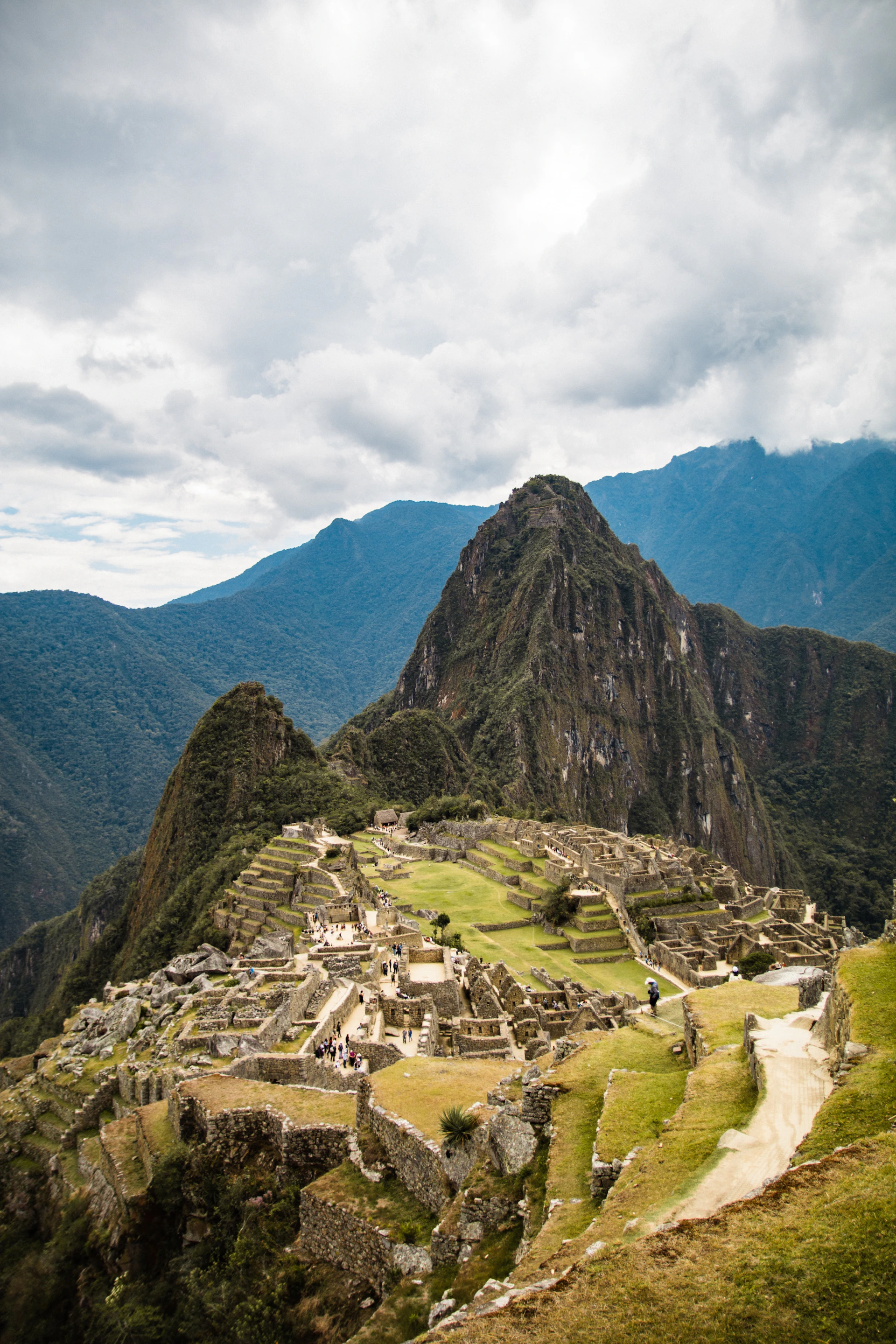 Ancient Incan ruins of Machu Picchu with terraces and stone structures, set against tall, green mountains under a cloudy sky.