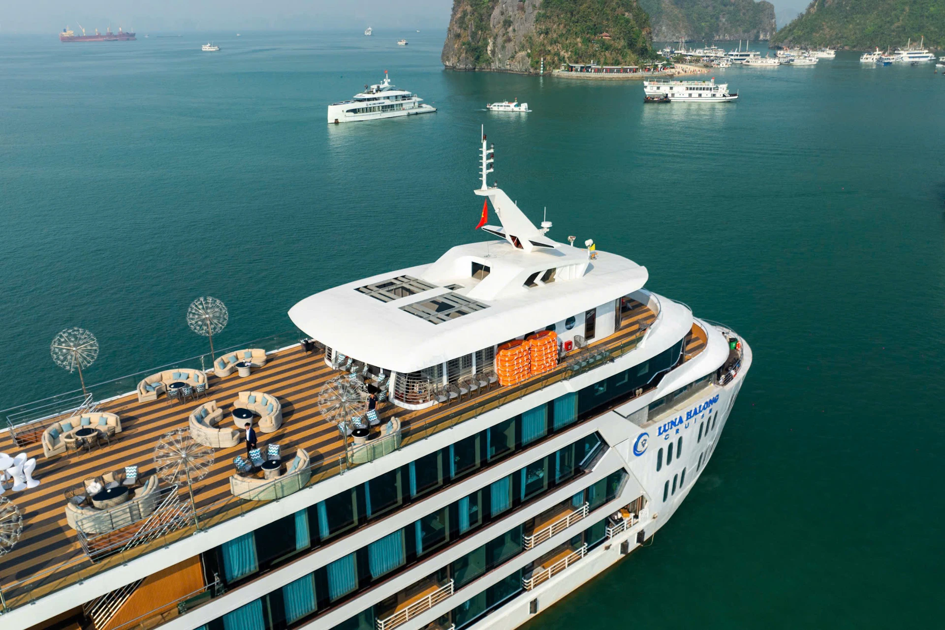 Large cruise ship docked in a scenic bay with smaller boats and island cliffs in the background.