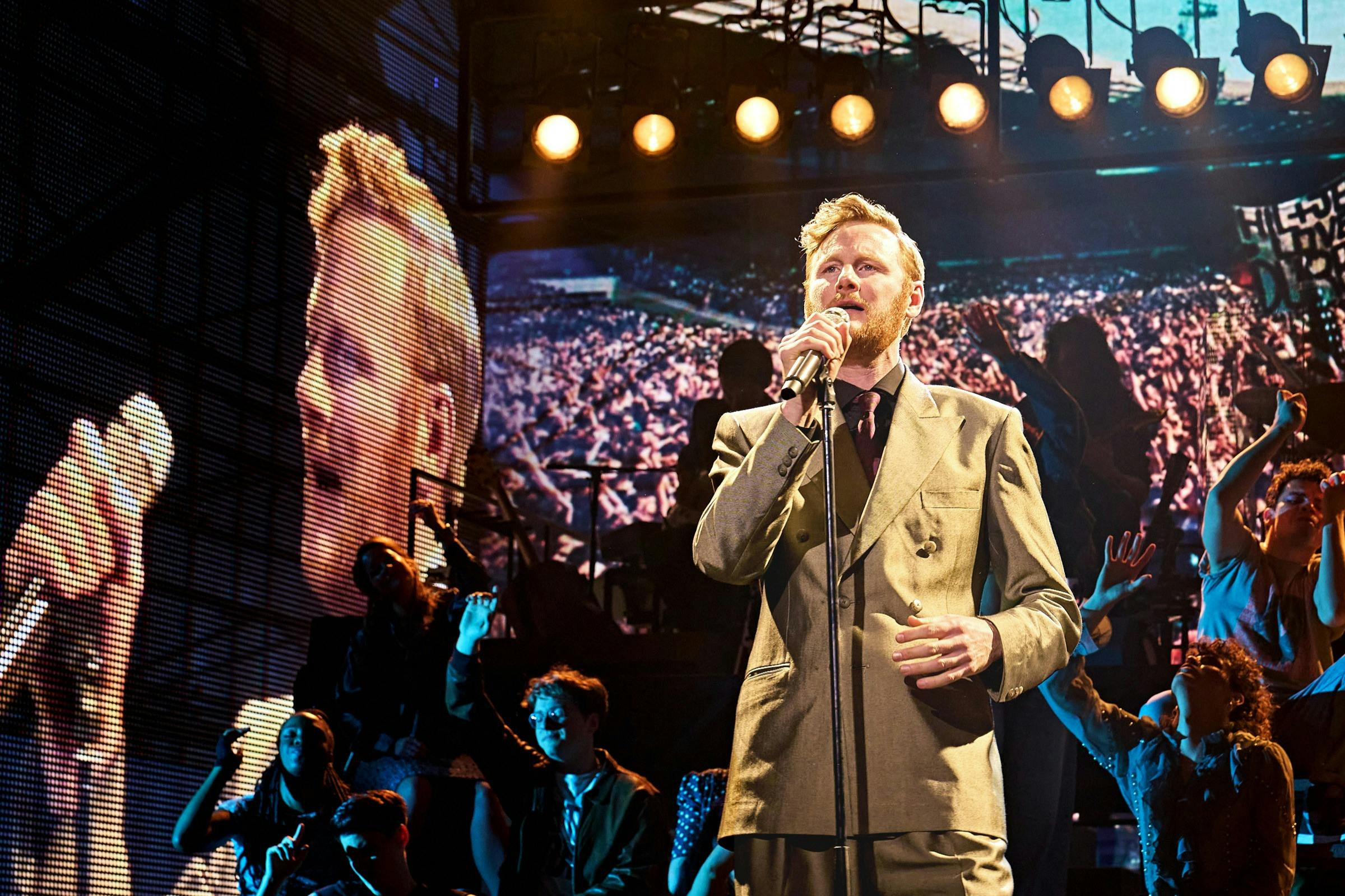 一名身穿西装的男子在舞台上对着麦克风唱歌，身后的屏幕上有一大群人，周围的人都围着他。