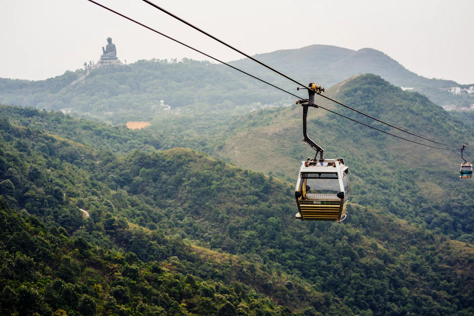 A cable car travels above a lush green hilly landscape with a large statue visible in the background.
