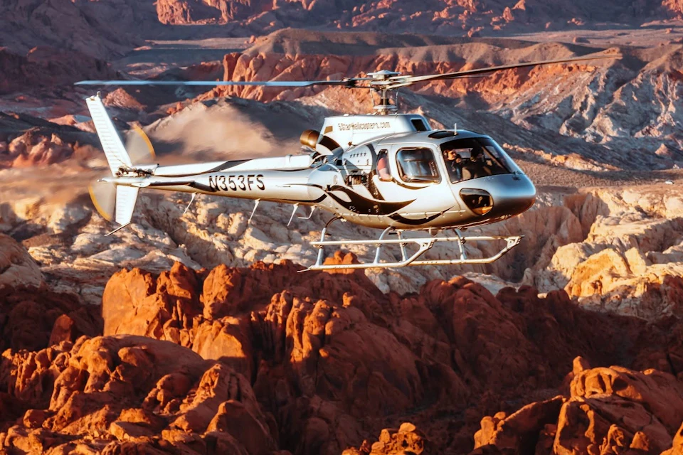 A helicopter flying over a scenic desert landscape with red rock formations and rugged terrain in the background.
