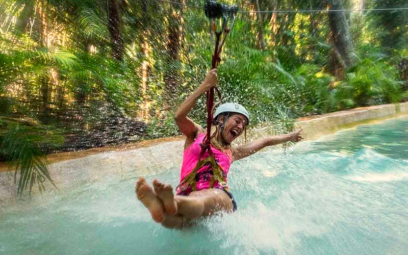 ピンクのシャツとヘルメットを着た女性は、緑の森の背景を持つ水にジップライニングをしています。