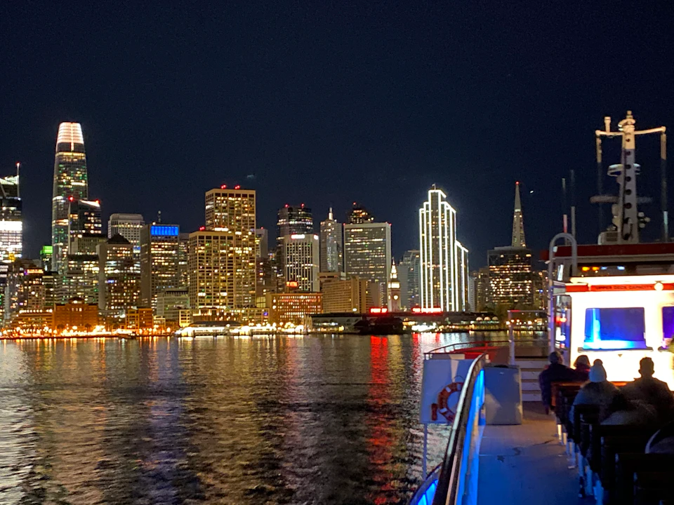 Suspension bridge silhouetted against an orange sunset with city skyscrapers in the background and water in the foreground.