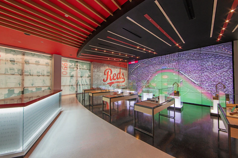 Modern museum exhibit featuring baseball memorabilia, a large "Reds" sign, and a giant screen displaying a crowded stadium.