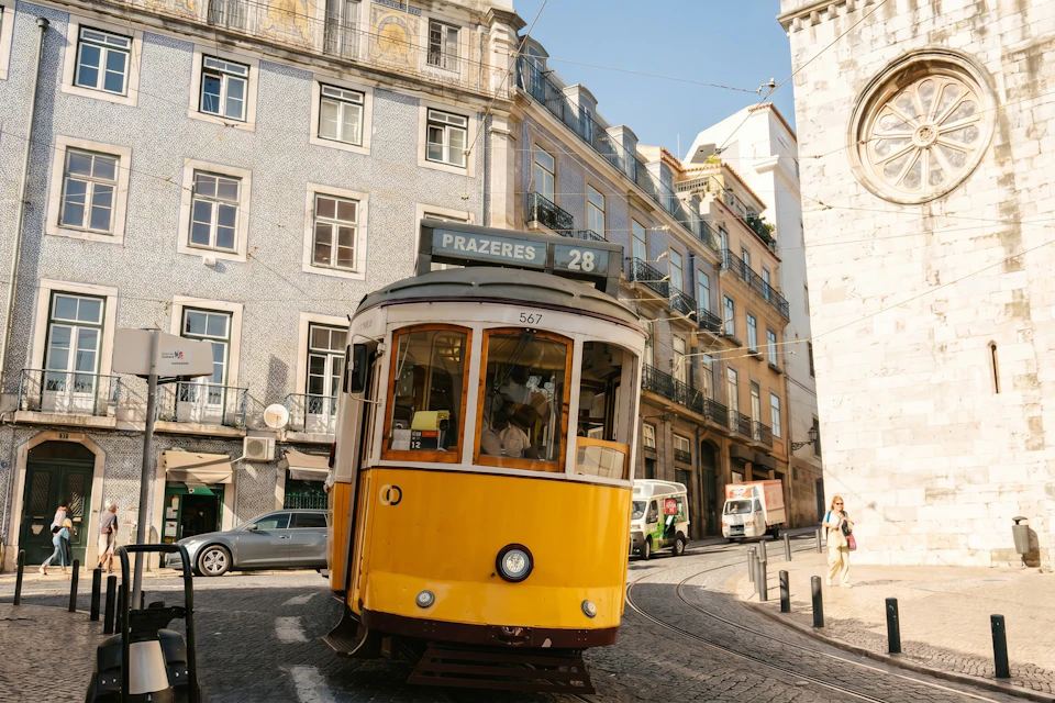 A yellow tram marked '28 Prazeres' on a street in a European city with historic buildings and a cobblestone road.
