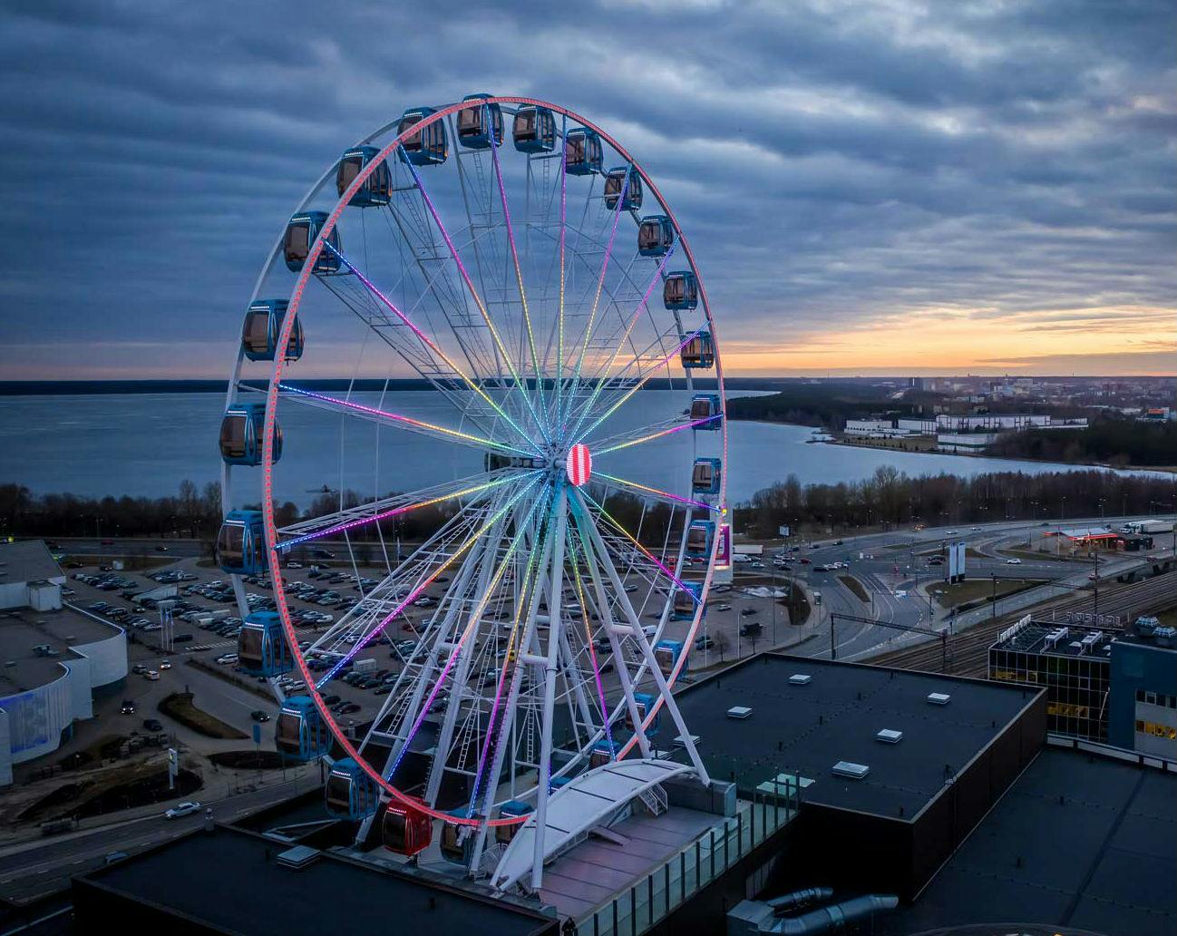 A large Ferris wheel with colorful lights stands near a waterfront at sunset with a cloudy sky and a parking lot below.