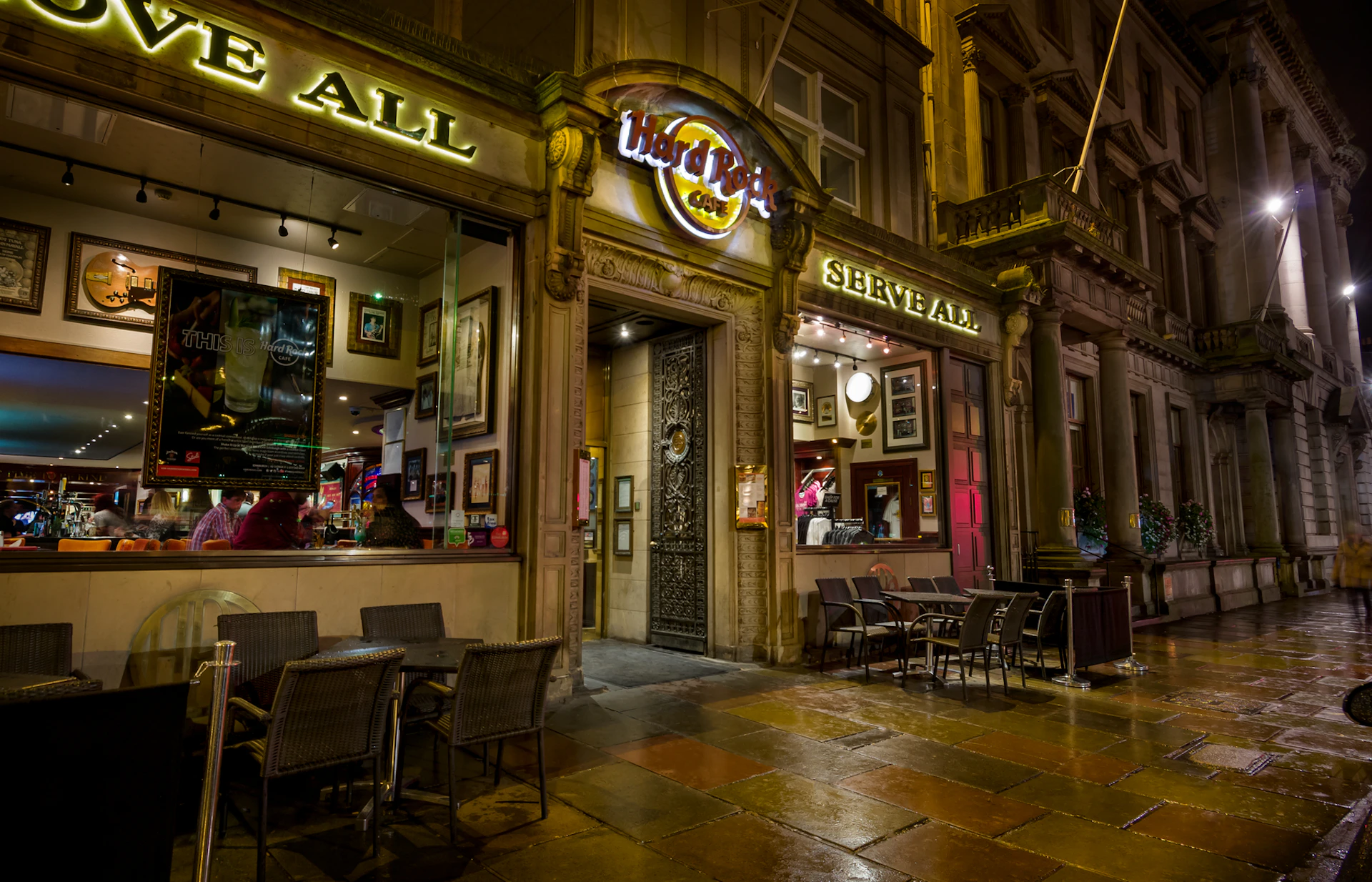 Entrance of a Hard Rock Cafe at night, with visible seating outside, illuminated signage, and framed pictures on the interior walls.