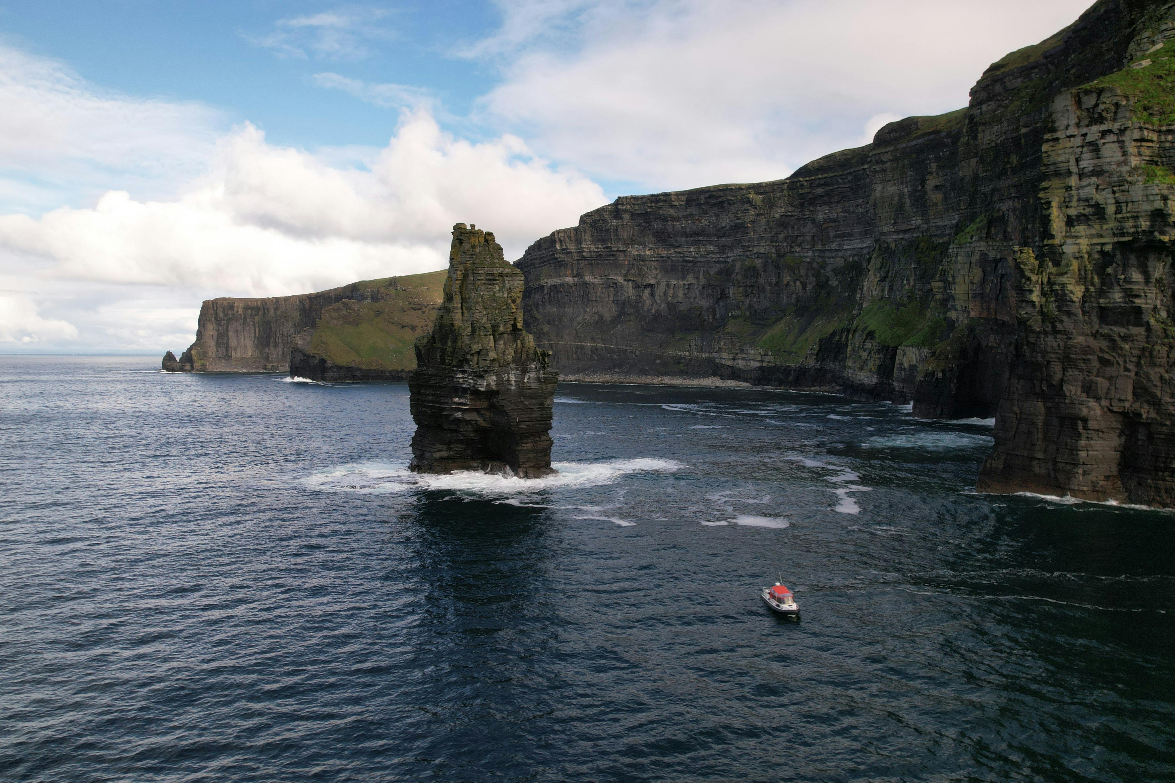 A sea stack near steep cliffs with a small boat on the water under a cloudy sky.