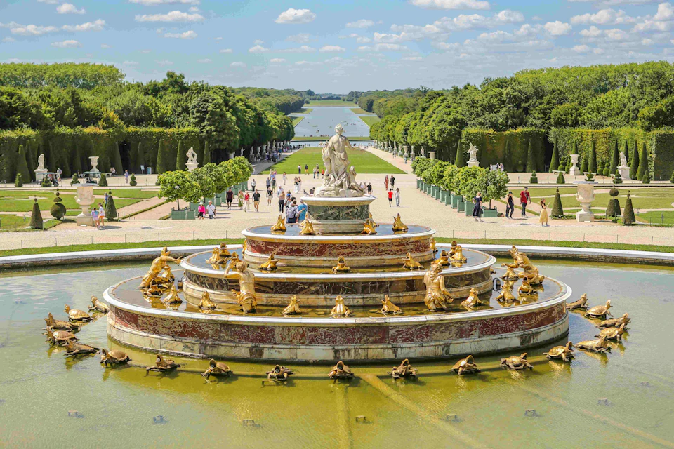 Grand fountain adorned with golden statues, people walking nearby, and a long canal surrounded by greenery in the background.