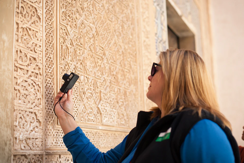 Person examining intricate wall carvings with a handheld device, wearing sunglasses and a vest, in an outdoor setting.