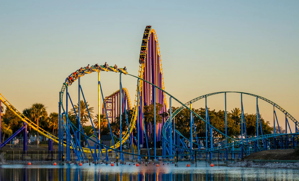 A colorful roller coaster with people riding is set against a backdrop of trees and water during sunset.