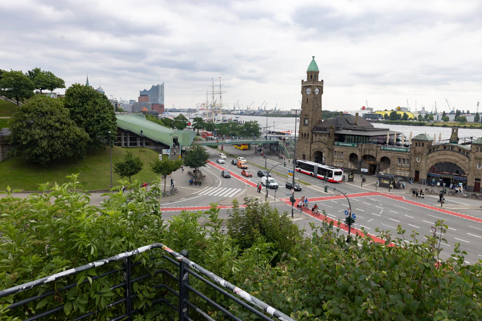 Urban scene with people, cars, a clock tower, and greenery. A harbor with ships and buildings in the background under a cloudy sky.