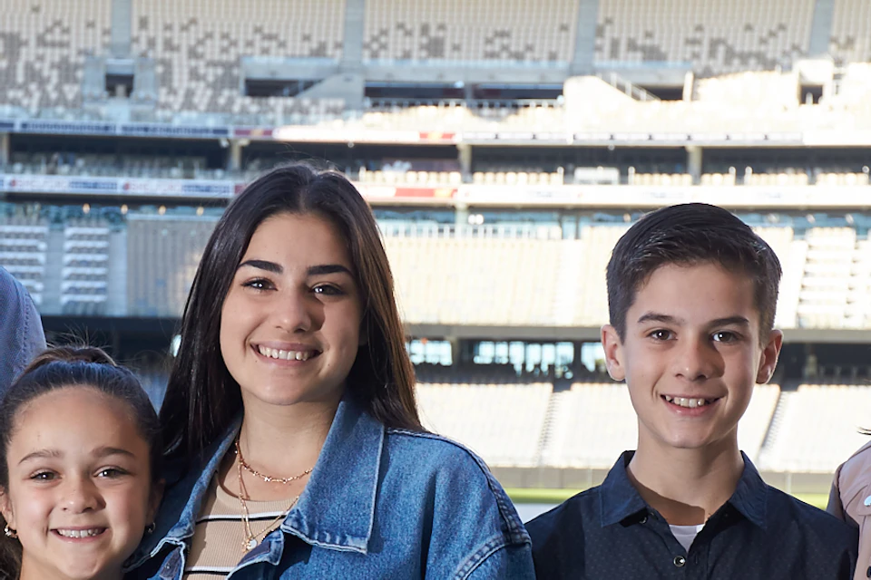 Five people stand smiling on a grassy field inside Optus Stadium, with empty stands in the background.