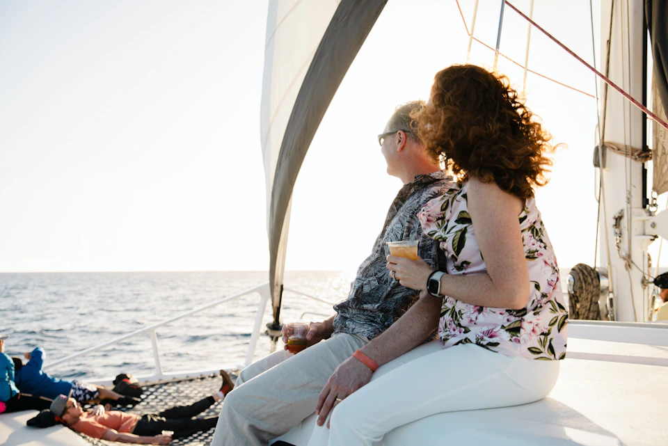 A couple sitting on a sailboat, holding drinks, gazing at the ocean under a bright sky. The boat's sail is visible.