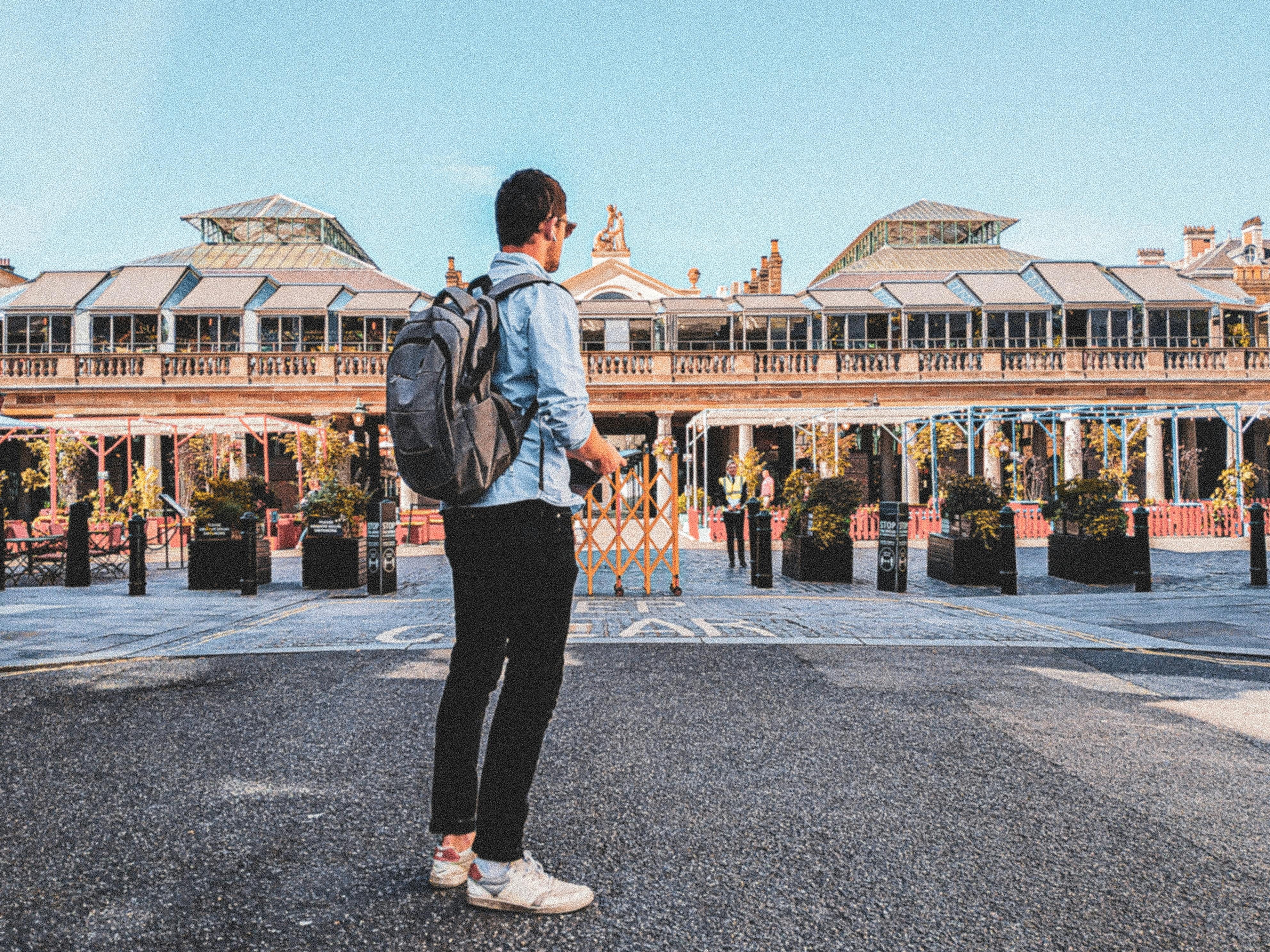 A person with a backpack stands on a street facing a large building with glass roof structures on a clear day.