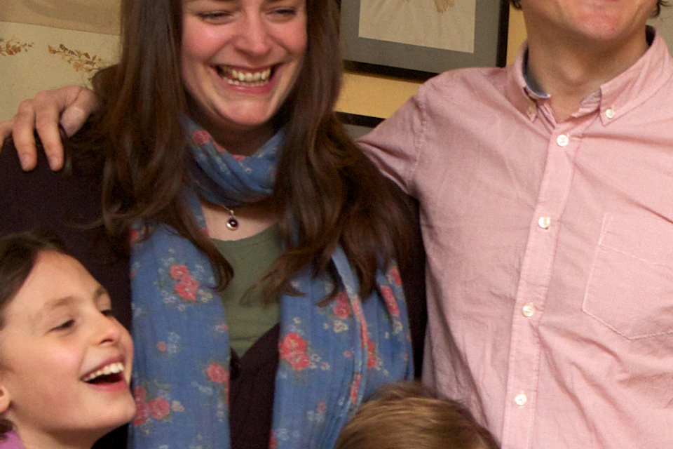 A smiling family of four stands in a room with framed pictures, a wooden table, and shelves displaying ceramics.