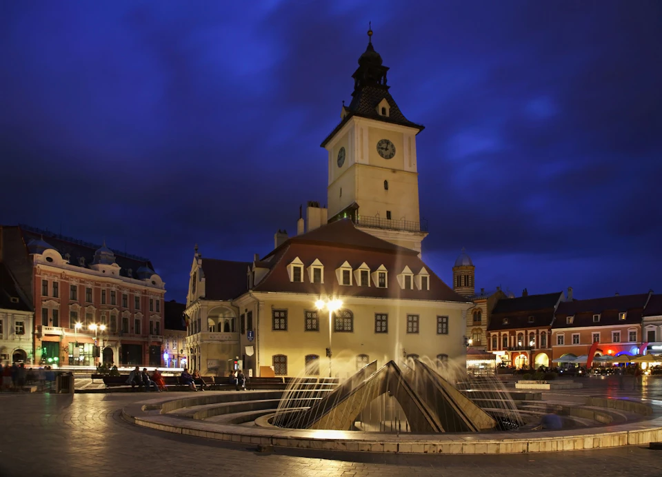 Night view of a clock tower and fountain in a square, surrounded by historic buildings with lit windows.