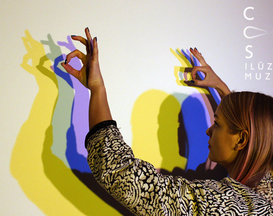 Person creating colorful hand shadows on a white wall, with overlapping yellow, blue, and purple light projections.