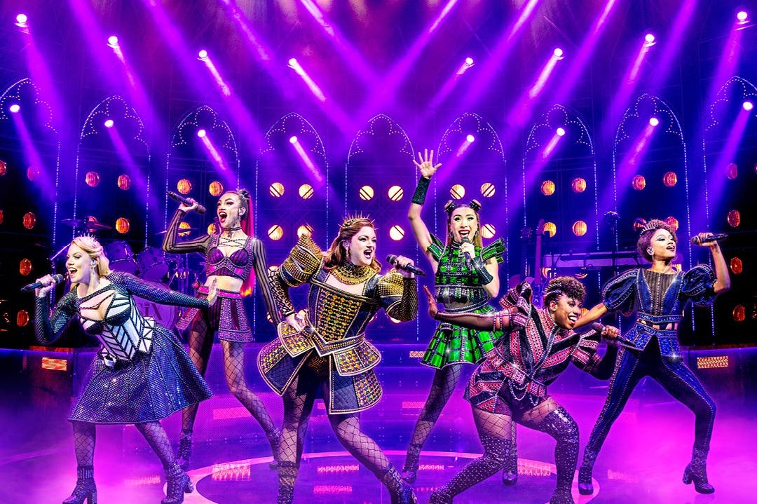 Artister på scenen i livliga kostymer som sjunger under lila och gul belysning, med en livlig bakgrund och kuliss.