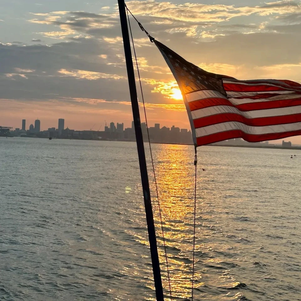 A U.S. flag waves on a boat at sunset. The sun reflects off the water, with a city skyline visible in the distance.