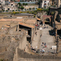 Ruins of an ancient city with stone structures, people walking along a wide pathway, and modern buildings and greenery in the background.