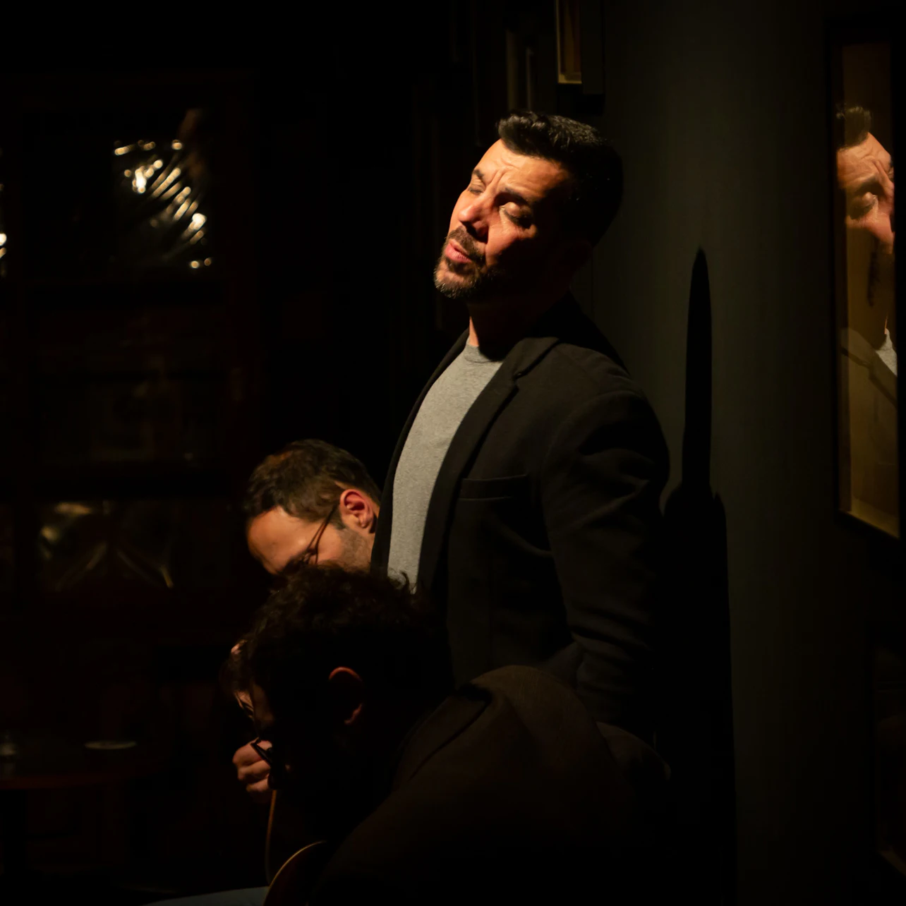 A man in a dark blazer stands against a dimly lit wall with a reflective surface, another man partially visible behind him.
