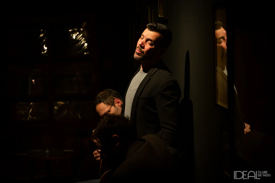 A man in a dark blazer stands against a dimly lit wall with a reflective surface, another man partially visible behind him.