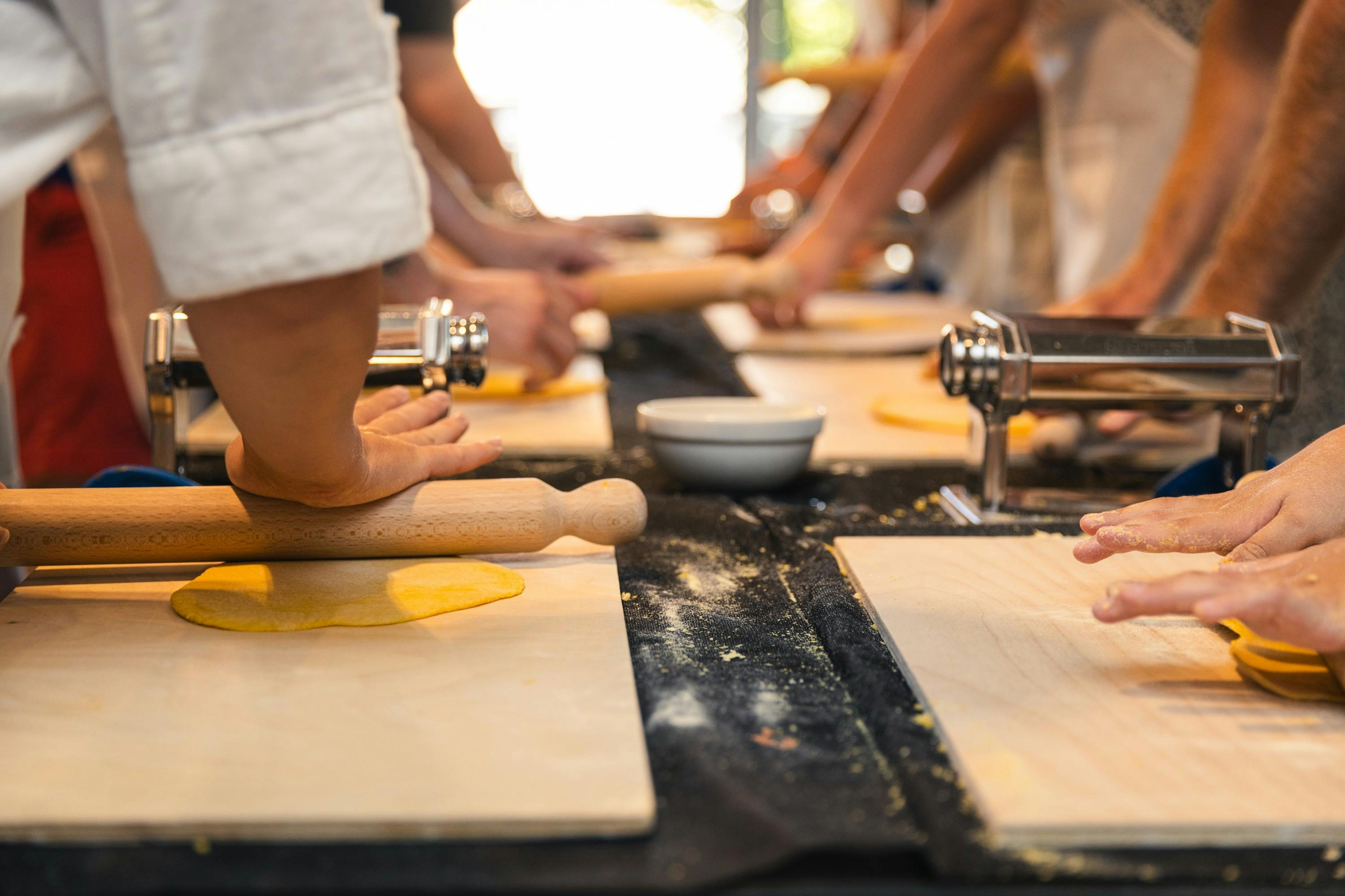 People rolling out dough on wooden boards with rolling pins and pasta machines on a table in a bright kitchen setting.