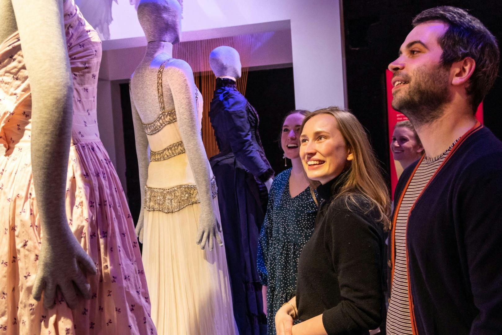 Eine Gruppe von Menschen, die lächelnd die ausgestellten Schaufensterpuppen in eleganten Outfits in einer musealen Umgebung betrachten.