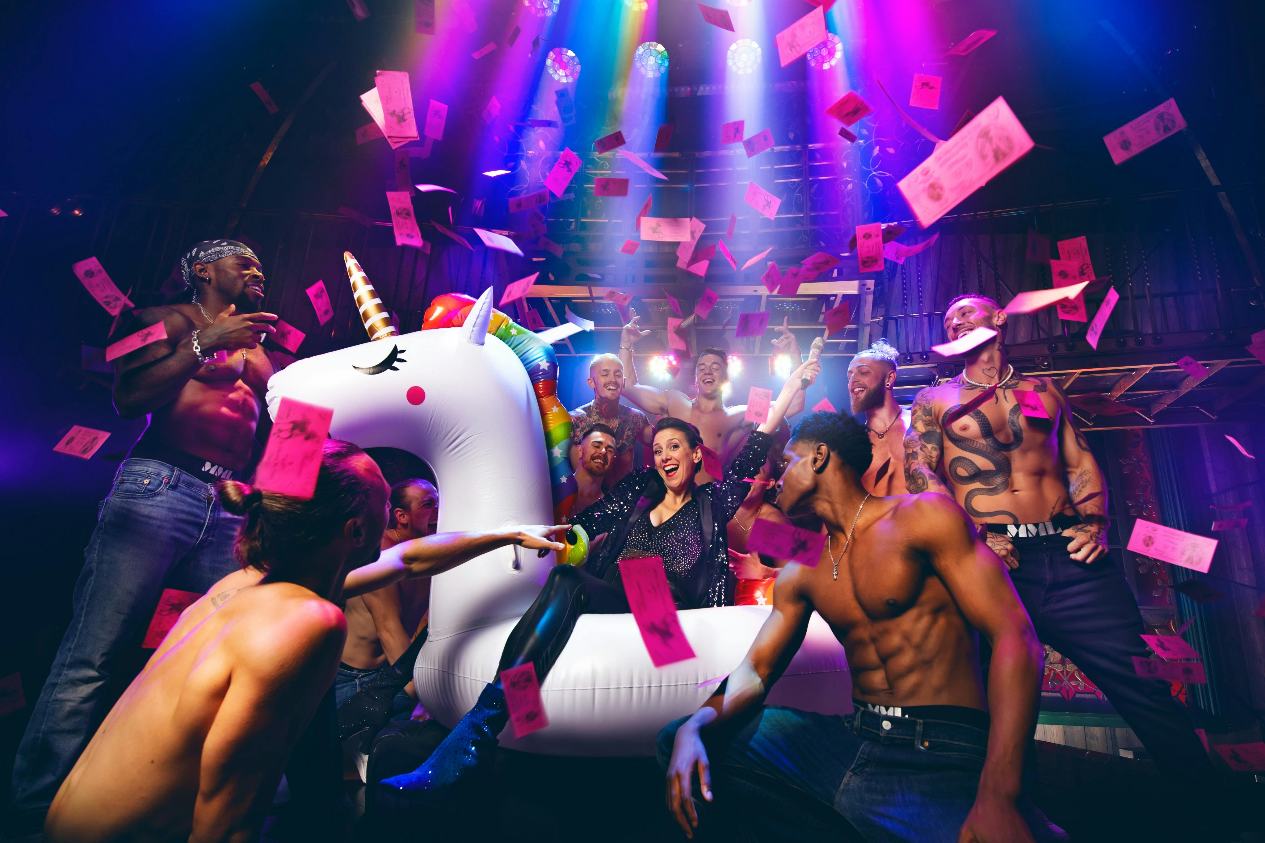 한 사람이 셔츠가 없는 사람들에 둘러싸인 팽창식 유니콘 위에 앉아 있고, 분홍색 종이 노트가 화려한 무대 조명 아래 있습니다.