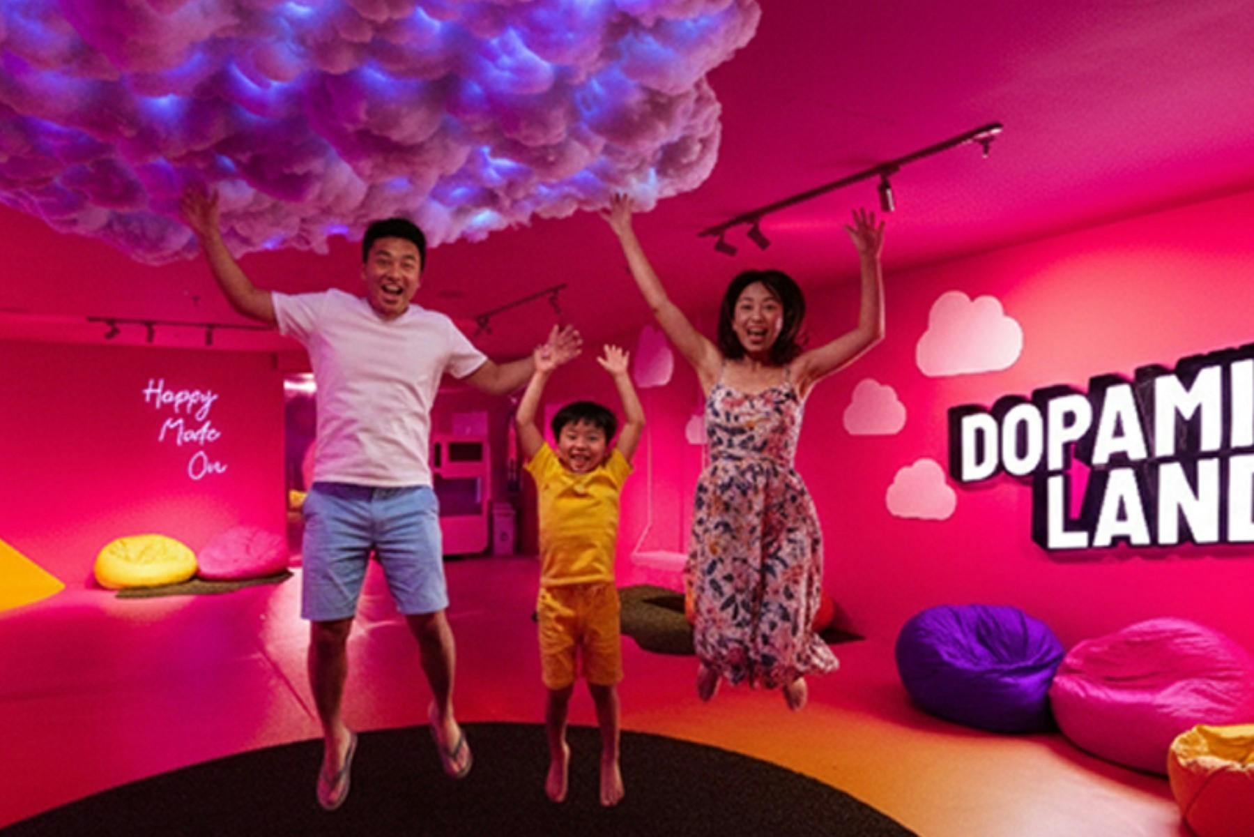 A family of three energetically jumping in an indoor space with pink lighting, cloud decorations, and playful decor.