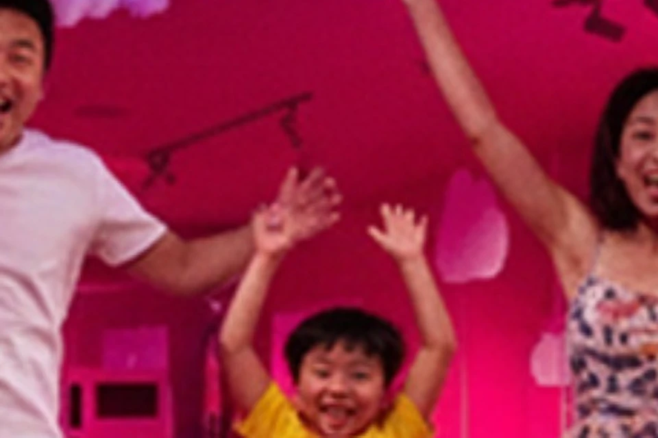 A family of three energetically jumping in an indoor space with pink lighting, cloud decorations, and playful decor.