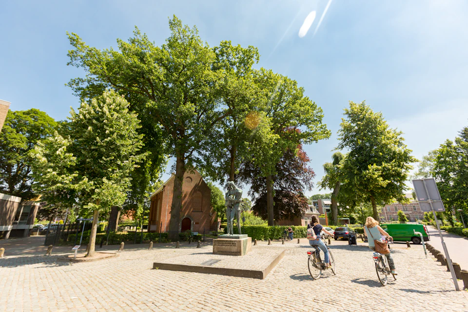 Cyclists ride near a statue in a sunny, tree-lined square with a brick building and cobblestone ground.