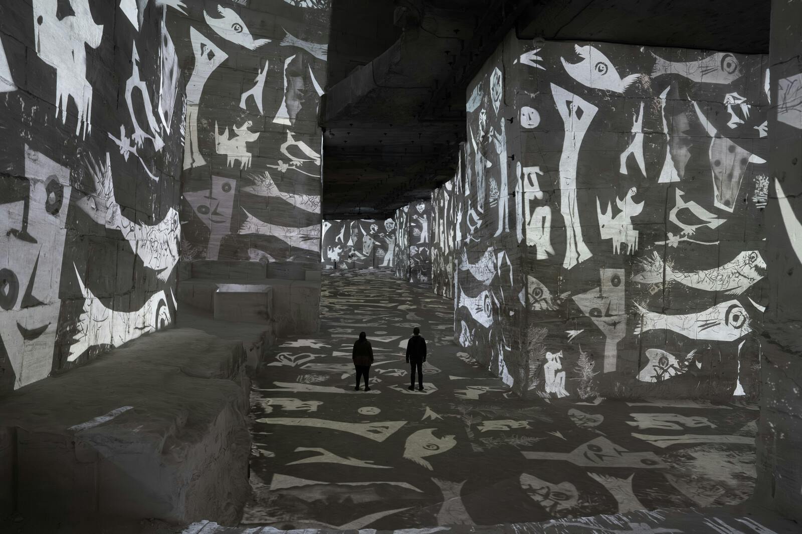 Two people walk in a dimly lit space with abstract black-and-white patterns projected on walls and floor.