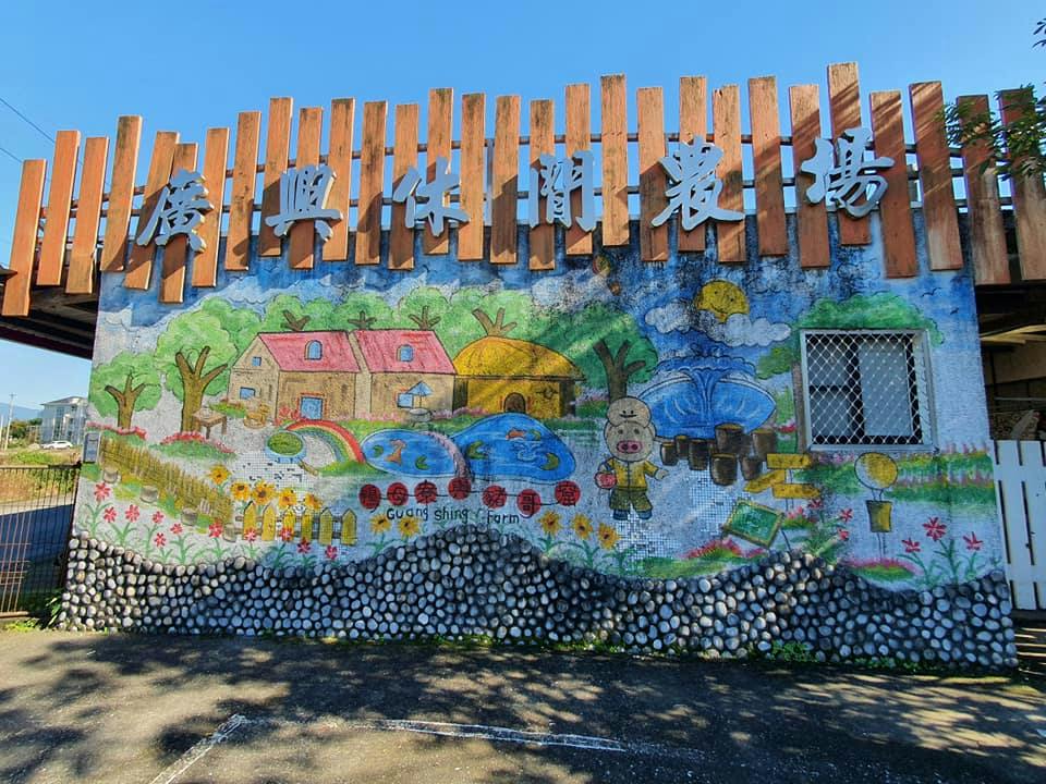 A colorful mural of a farm scene with houses, fields, and animals on a stone wall, topped with wooden slats and Chinese text.