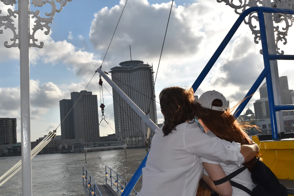 Two people stand on a boat, looking at a cityscape with tall buildings across the water under a partly cloudy sky.