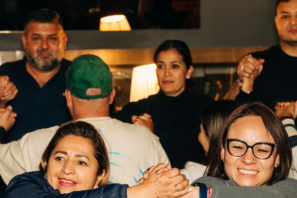 A group of people in a room, holding hands in a circle, smiling and engaging in an interactive activity.